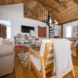 A cozy interior of a small cabin with a rustic wooden aesthetic. The room features a white paneled wall on the right with a flat-screen TV mounted above a white bookshelf filled with various books and knick-knacks. There's a comfy armchair with a zigzag-patterned throw blanket in the foreground, and a wooden ladder leading to a lofted area above.